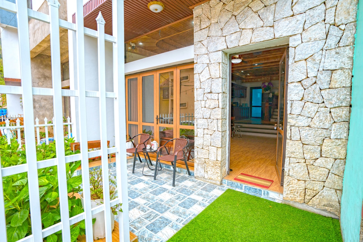 The entrance features a stone facade with a wooden door, leading to a charming outdoor terrace furnished with two chairs. A patterned stone walkway and greenery create a welcoming transition from the outdoors to the interior space.