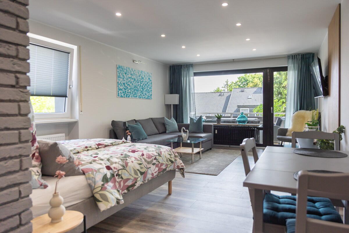 An open-plan living space features a comfortable double bed draped in floral bedding and a dark gray sofa against a light wall. A dining table is positioned near the large glass doors, leading to a balcony with greenery visible outside.
