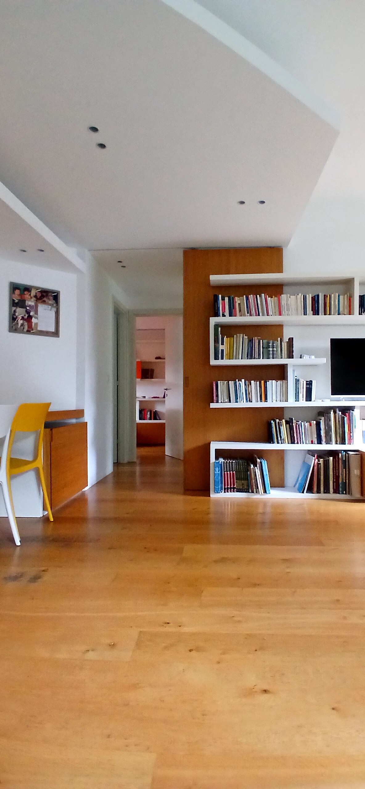 A spacious interior featuring light wooden flooring and built-in bookshelves filled with books. A sleek, modern desk with a yellow chair is positioned in the corner, while a doorway leads to the kitchen area visible in the background.