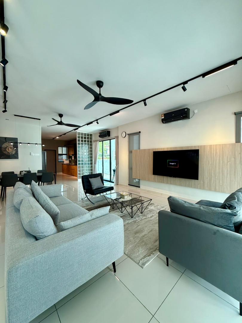 A modern living area is equipped with comfortable gray sofas, a glass coffee table, and a sleek wall-mounted TV. Natural light is filtered through large windows, while the open layout connects to the dining area in the background. Ceiling fans contribute to the airy ambiance.
