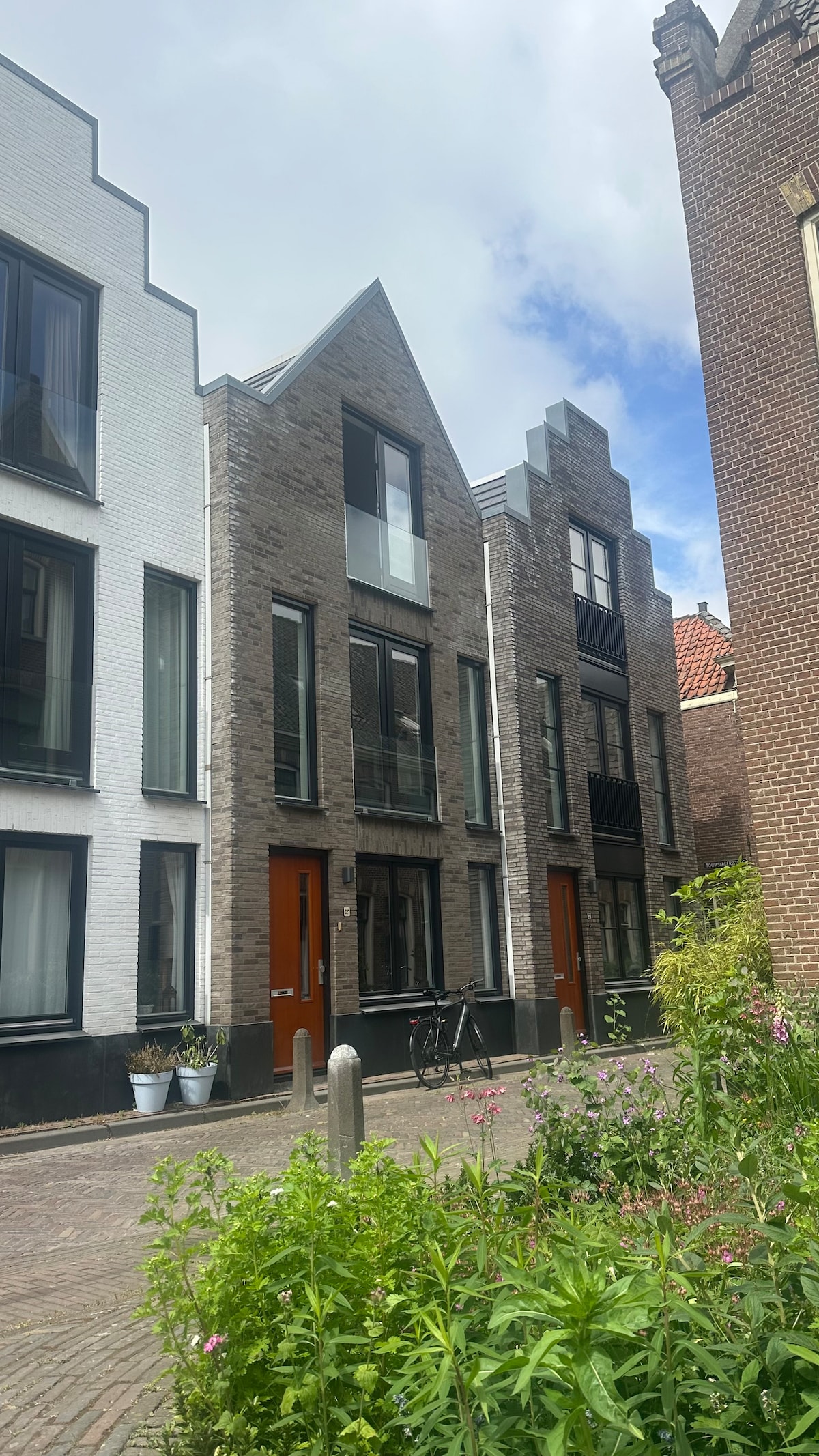 A modern building is showcased, featuring a combination of brick and white exterior. Large windows allow for natural light, and wooden doors provide an inviting entrance. Lush greenery and flowers are visible in the foreground, creating a vibrant urban atmosphere.