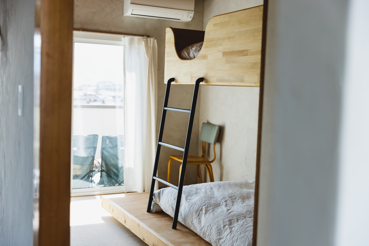 A compact guest room features a unique two-tiered bed, showcasing smooth wooden materials. A ladder provides access to the upper sleeping area. Soft natural light enters through large glass doors leading to a balcony, where two chairs are positioned for outdoor seating.