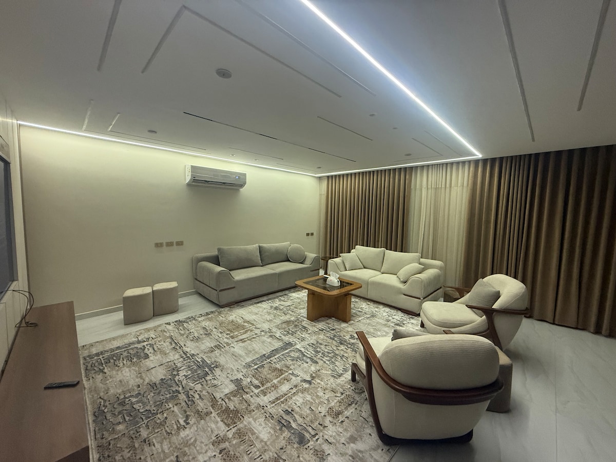 Een luxe woonkamer met een elegant, modern design, comfortabele meubels in een rustige beige kleur en een plafond met geometrische ledverlichting die een verfijnde artistieke uitstraling toevoegt. De marmeren vloer en het luxe tapijt versterken de luxe van de plek. Perfect voor comfort en ontvangst