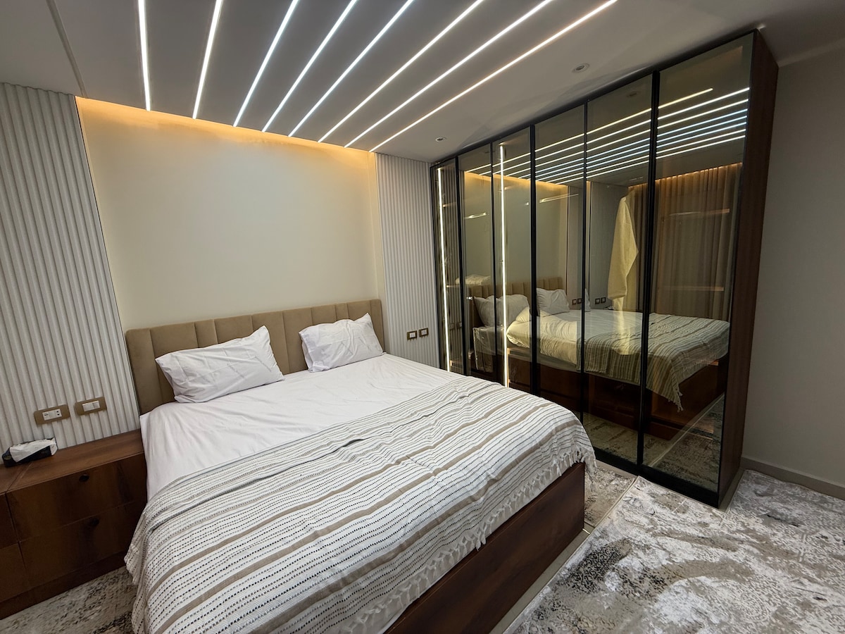Een elegante hoofdslaapkamer met een moderne inrichting, een groot bed en elegante spiegelkasten, met led-plafondverlichting die een luxe tintje toevoegt. De vloer is bedekt met luxe tapijt en zware gordijnen voor volledige privacy. De kamer heeft een eigen badkamer voor meer comfort