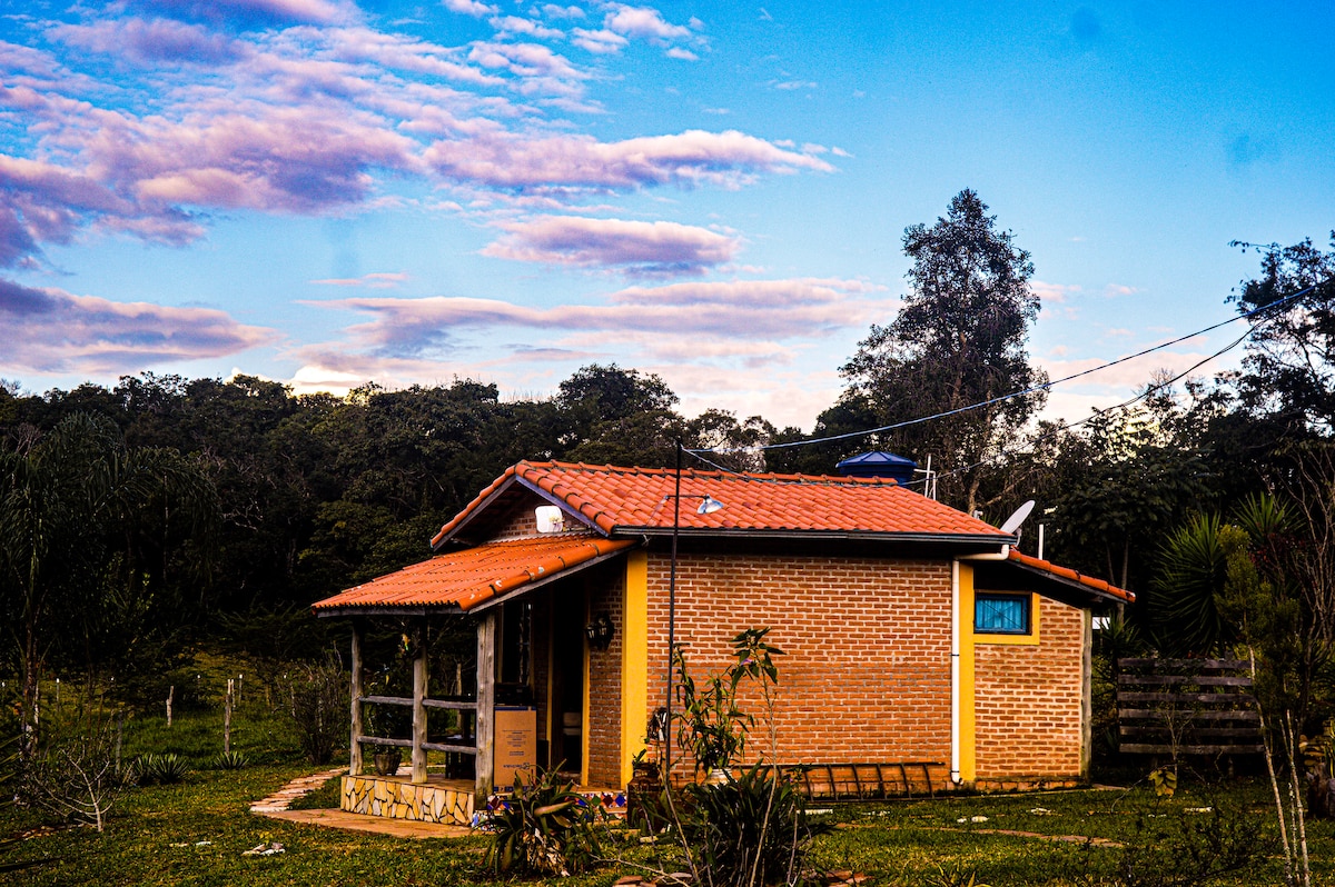 A charming chalet is positioned amidst a serene natural setting, featuring a terracotta roof and warm brick exterior. Surrounding greenery and trees enhance the tranquil atmosphere, while a clear blue sky with soft clouds adds to the peaceful scenery.