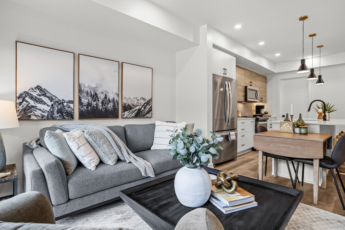 A cozy living area features a plush gray couch adorned with soft throw blankets. Scenic mountain artwork is displayed on the wall, enhancing the ambiance. A modern dining table paired with sleek chairs, surrounded by warm lighting, complements the fully equipped kitchen visible in the background.