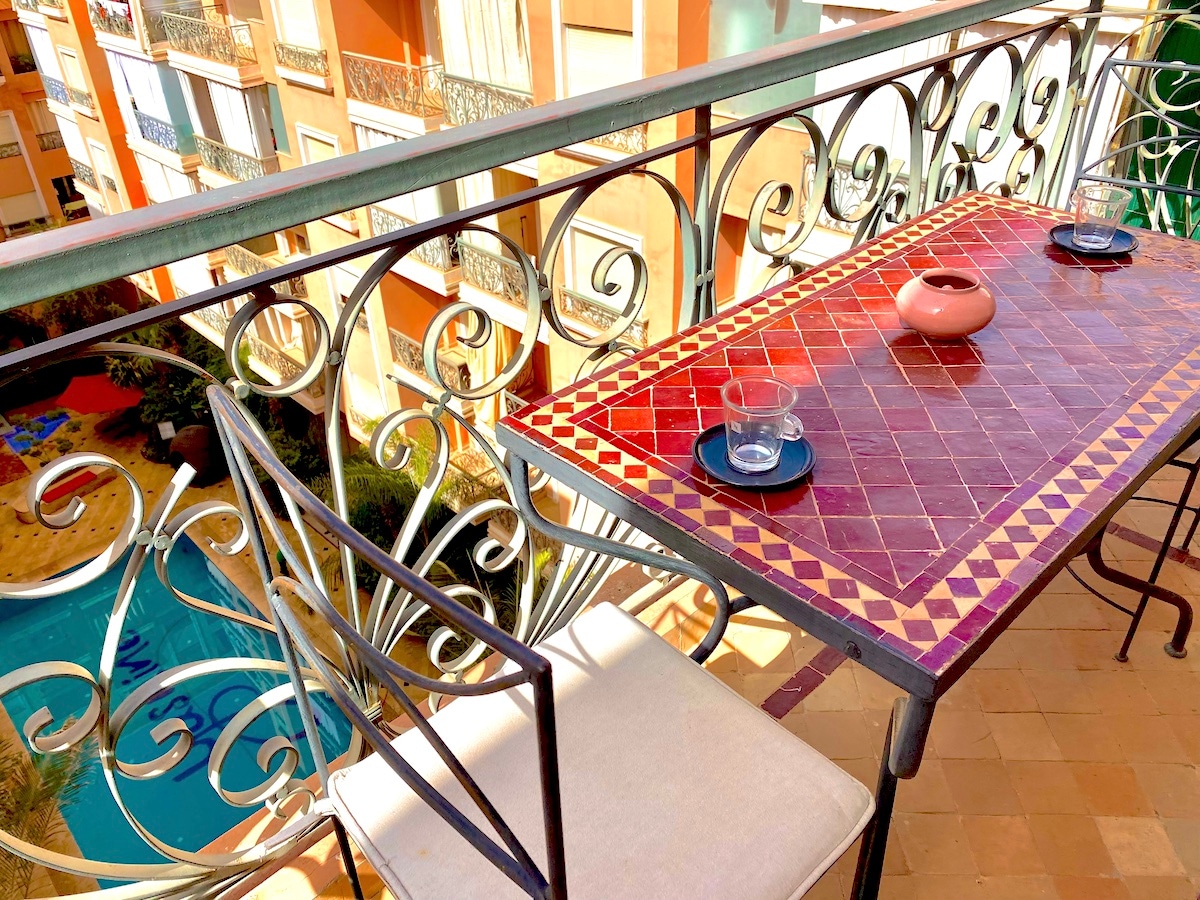 A decorative balcony table is set with two glasses and a small pot, overlooking a swimming pool. The table features a vibrant patterned surface, while wrought iron chairs provide comfortable seating. Natural light enhances the warm tones of the decor.
