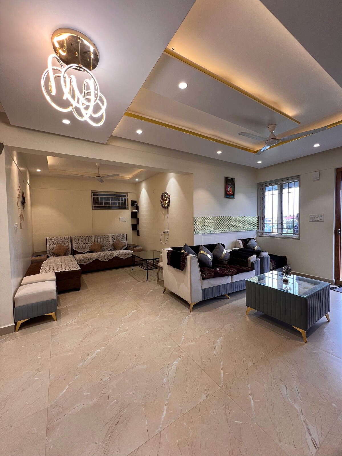 A modern living area is illuminated by stylish ceiling lights. Comfortable seating arrangements include a sectional sofa and armchairs, complemented by a glass coffee table. Large windows allow natural light to flow in, enhancing the spacious and welcoming ambiance.