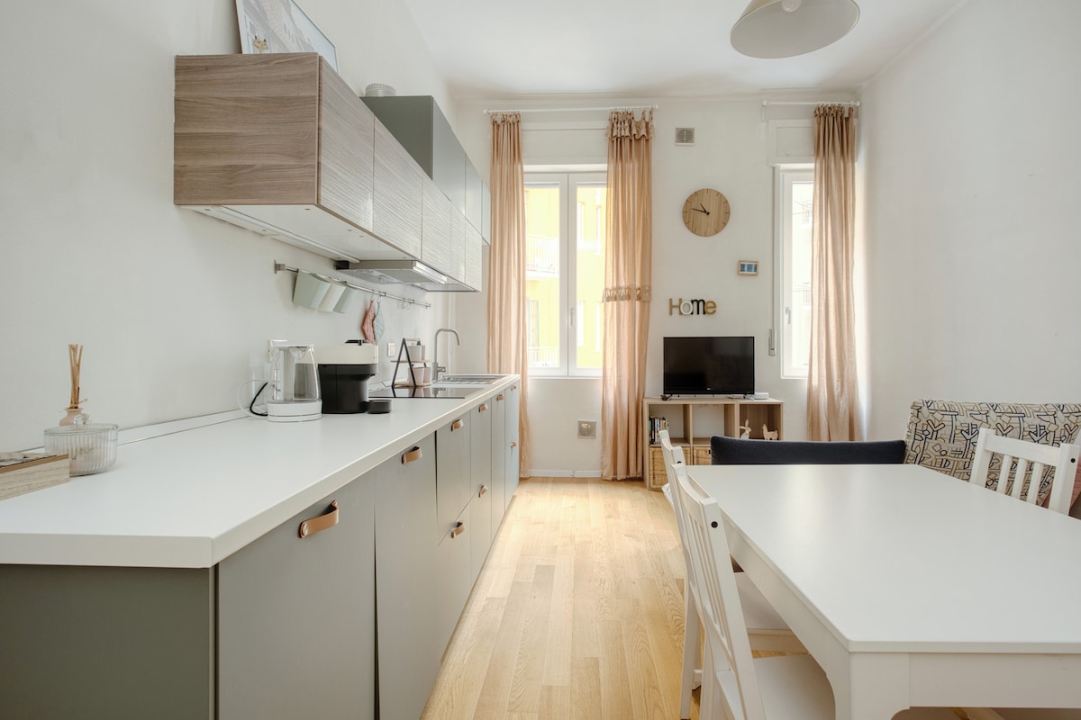 A bright kitchen area is equipped with modern cabinetry and a spacious countertop. A coffee maker and kettle are visible, alongside a furnished dining table and chairs. Natural light enters through large windows, and a clock is mounted on the wall, contributing to a functional atmosphere.