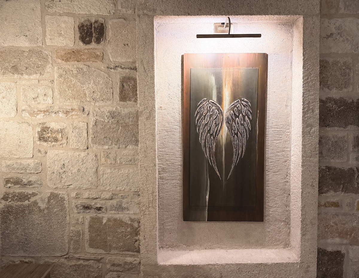 A piece of wall art featuring a pair of stylized wings is displayed in a softly lit alcove. The artwork is framed within a textured stone wall, creating a warm and inviting focal point in the space.