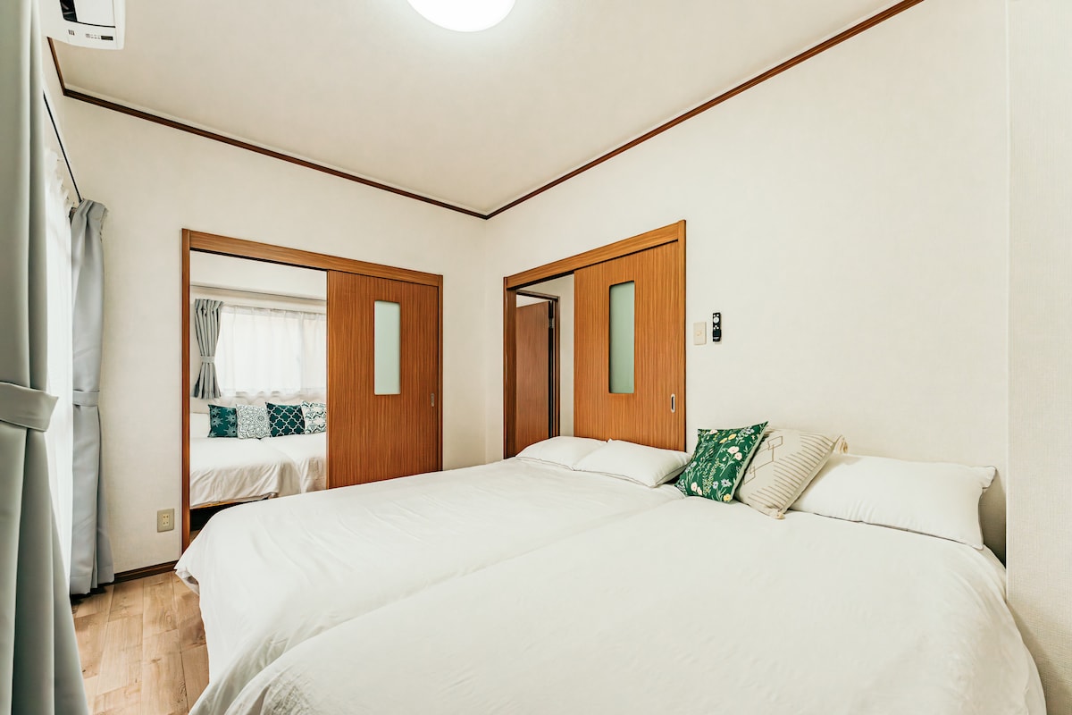 A well-lit bedroom features two spacious double beds arranged with a decorative cushion on one. Two doors lead to adjacent rooms, while curtains frame the windows, allowing natural light to illuminate the space.