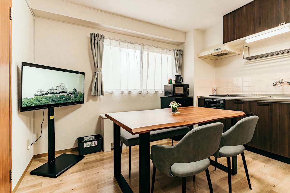 A dining area is presented with a wooden table accompanied by two upholstered chairs. A floor-mounted television displays an image of a castle. Natural light filters through a large window, accentuated by light-colored curtains. A kitchenette is visible in the background.