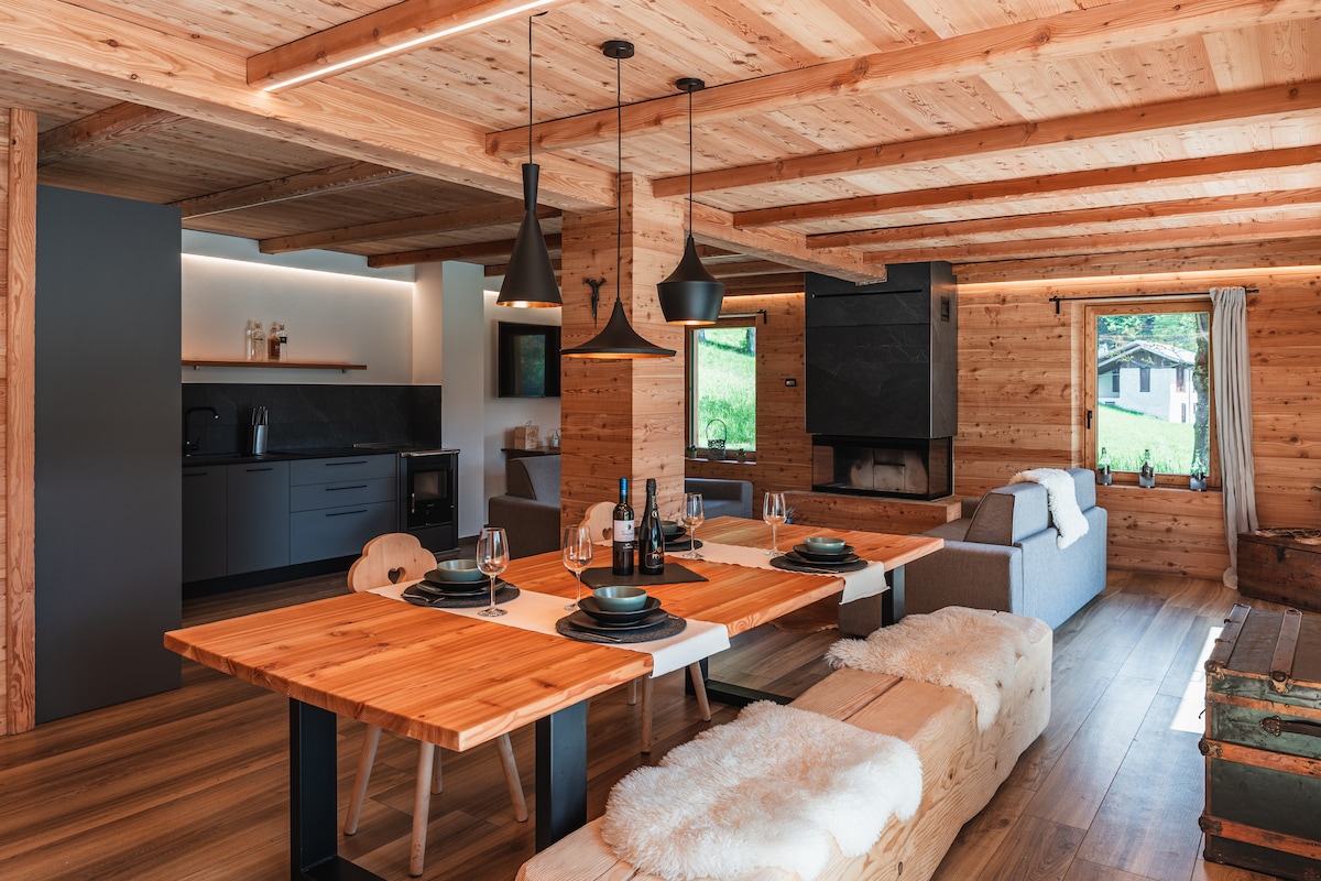 The spacious living area features a wooden dining table accompanied by black dining chairs and wooden benches. A modern kitchen with sleek cabinetry is visible, along with a cozy seating arrangement highlighted by a black fireplace. Natural light filters in through large windows.