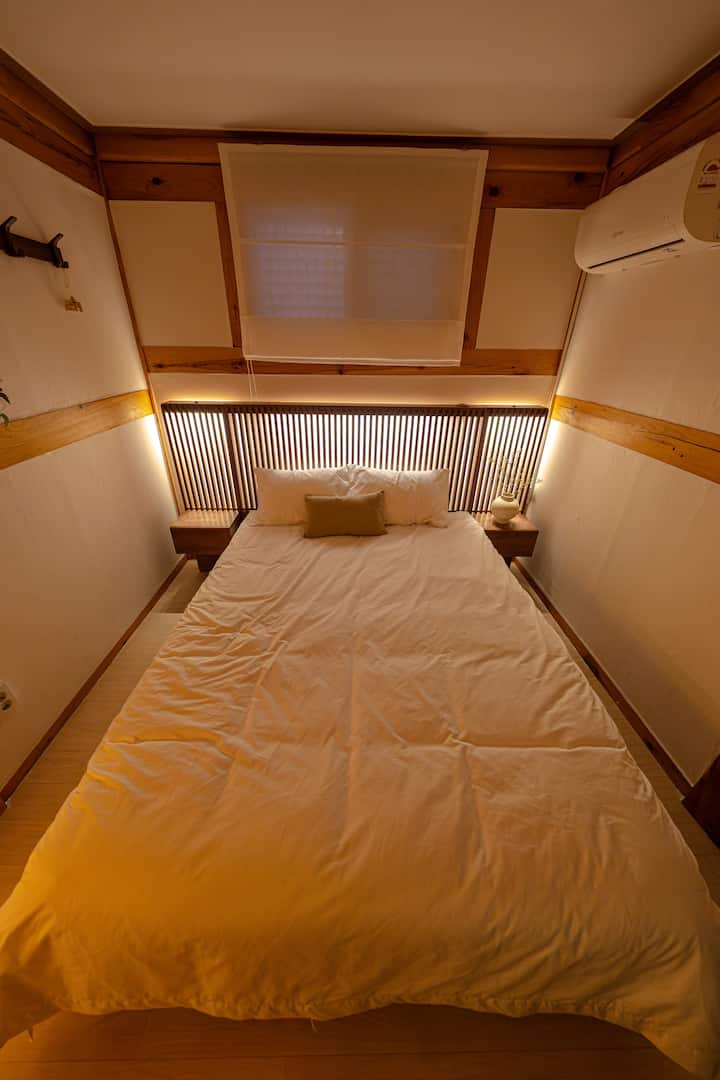 Спалня с деликатно осветление, което се разпространява по стените и създава уютна атмосфера. В края на деня топлото одеяло ще заглуши шума на града и ще успокои умствената ви тревожност. Смес от традиционните изчистени линии на ханока и модерните удобства правят това място оптимално убежище за добър сън. Спокойна нощ, вашата спокойна нощ започва тук.