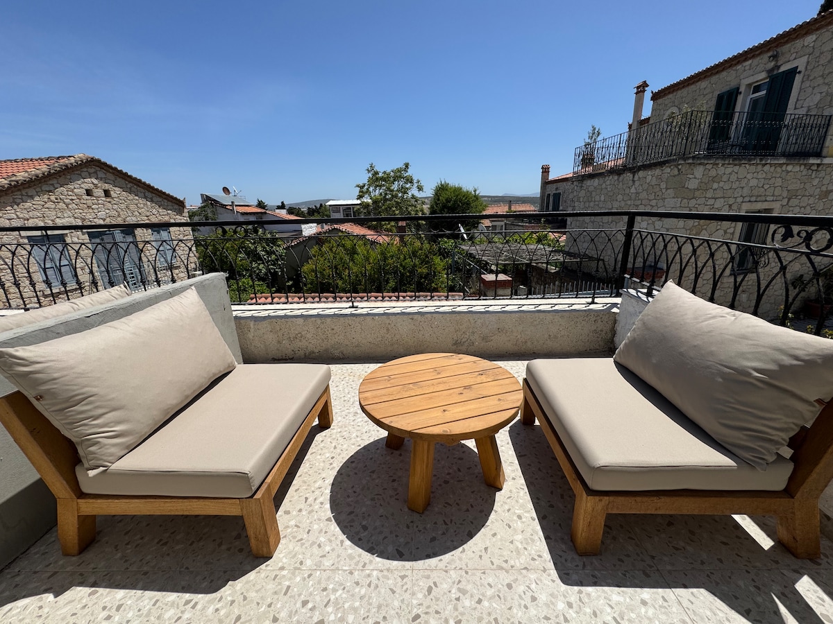 A private terrace is displayed, featuring two comfortable lounge chairs with soft cushions and a small wooden table in between. The background reveals a serene view of stone buildings and greenery under a clear blue sky.