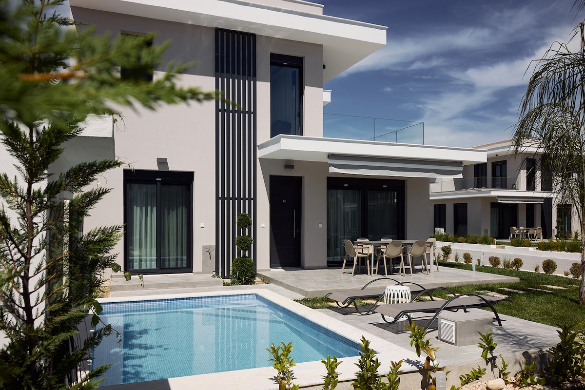 A modern two-story villa features a private pool surrounded by sunbeds, set against a backdrop of lush landscaping. The outdoor area includes a dining set, providing a space for relaxation and enjoyment. Large windows invite natural light into the interior.