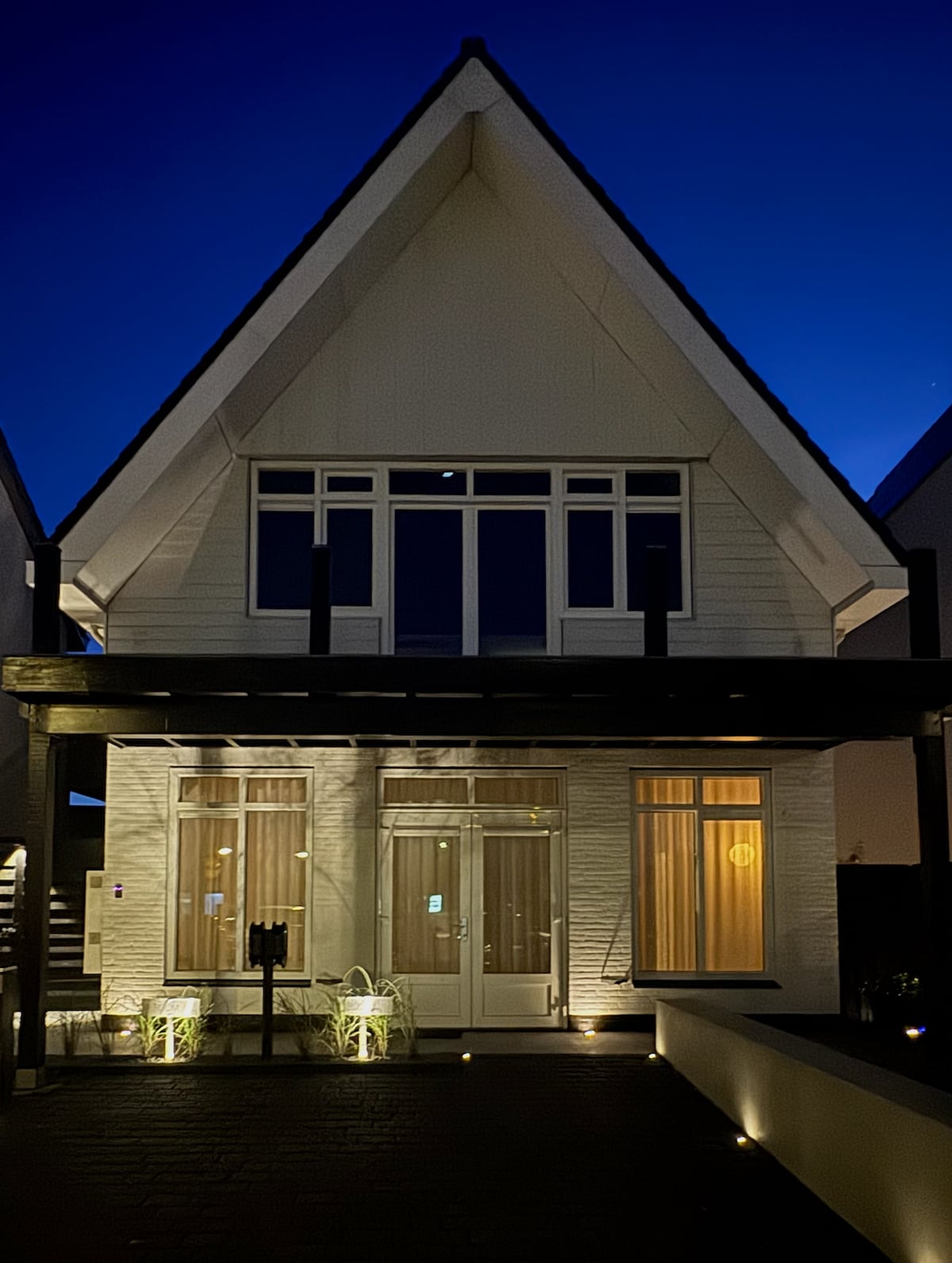A modern house is featured with a steep roof and large front windows. Soft outdoor lighting highlights the entrance, enhancing the architectural lines. The porch is bordered by minimalist landscaping, creating an inviting exterior that reflects a blend of contemporary design and a serene atmosphere.