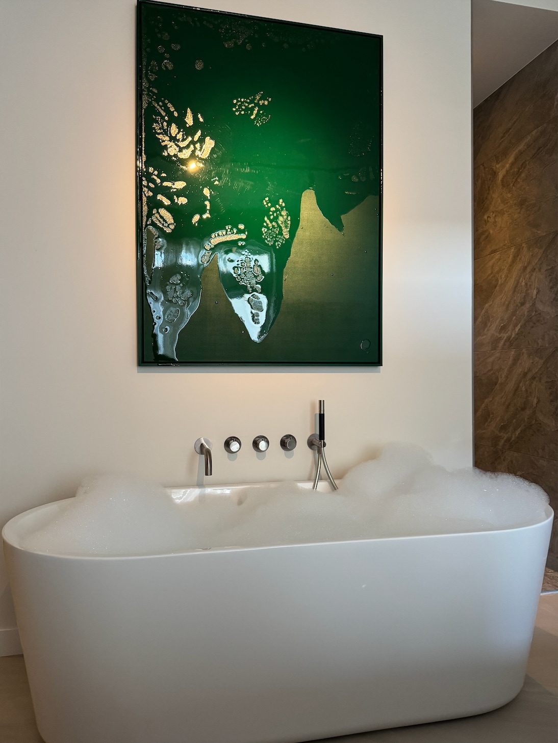 A modern freestanding bathtub is filled with bubbles, positioned against a wall featuring an artistic green piece. The tub is accompanied by sleek, contemporary fixtures that enhance the space's design, while soft lighting creates a calming environment.