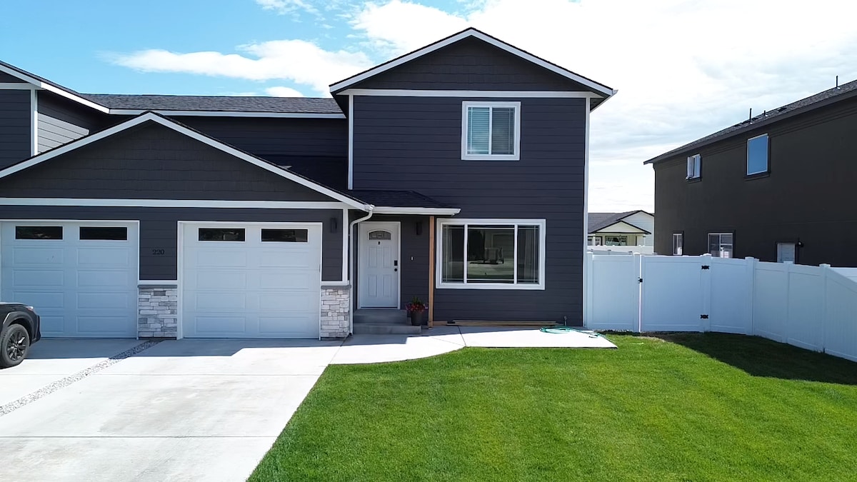 A modern two-story building with a gray exterior is presented. The front features a neatly trimmed lawn and a driveway leading to two garages. Large windows allow natural light in, while a small flower pot is placed near the entrance, bordered by a white fence.