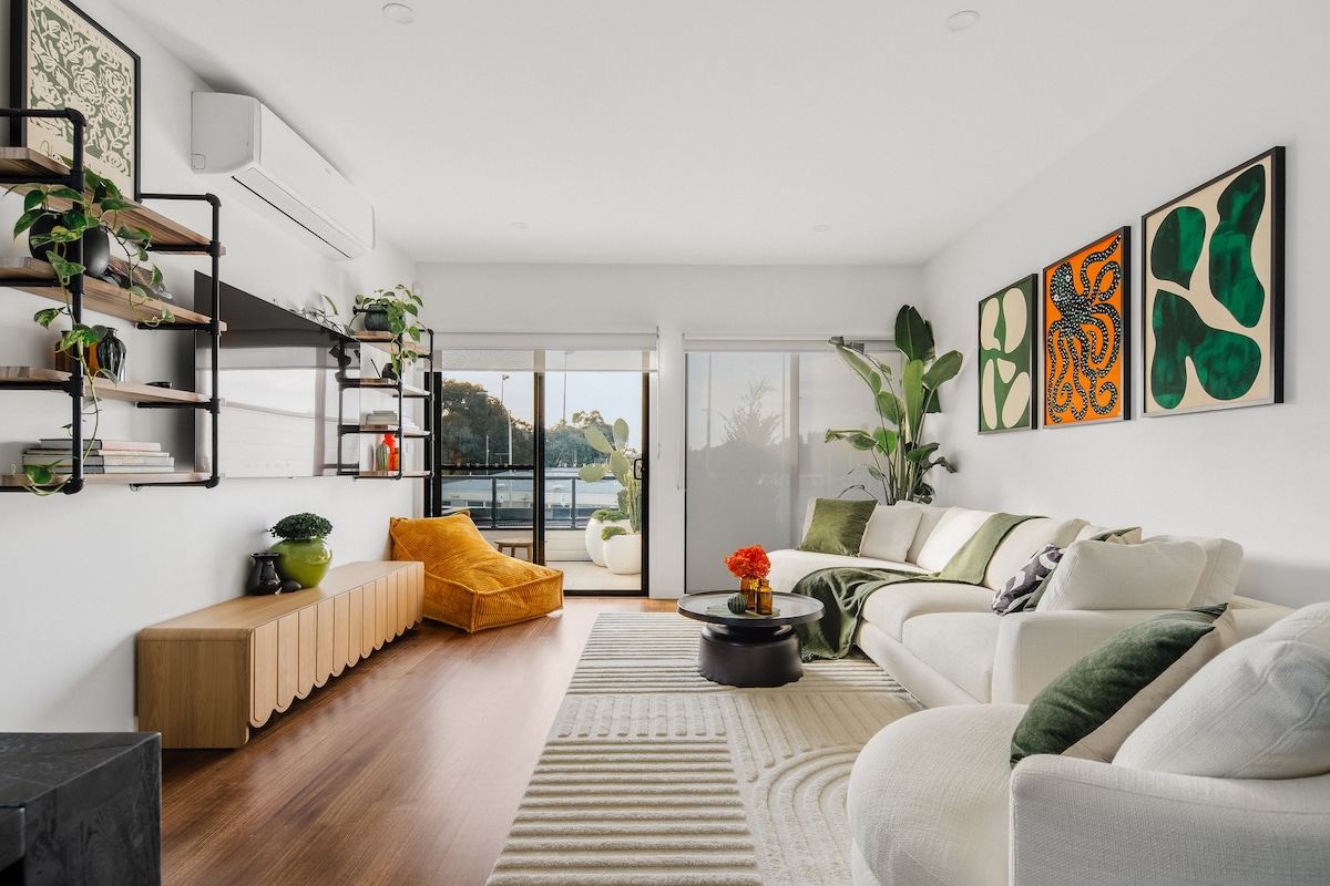 A sunlit living area showcases modern furnishings with a cozy white couch and a vibrant yellow chair. Green plants and framed artwork adorn the walls, while sliding glass doors provide access to outdoor views. A circular black coffee table sits on a textured rug.