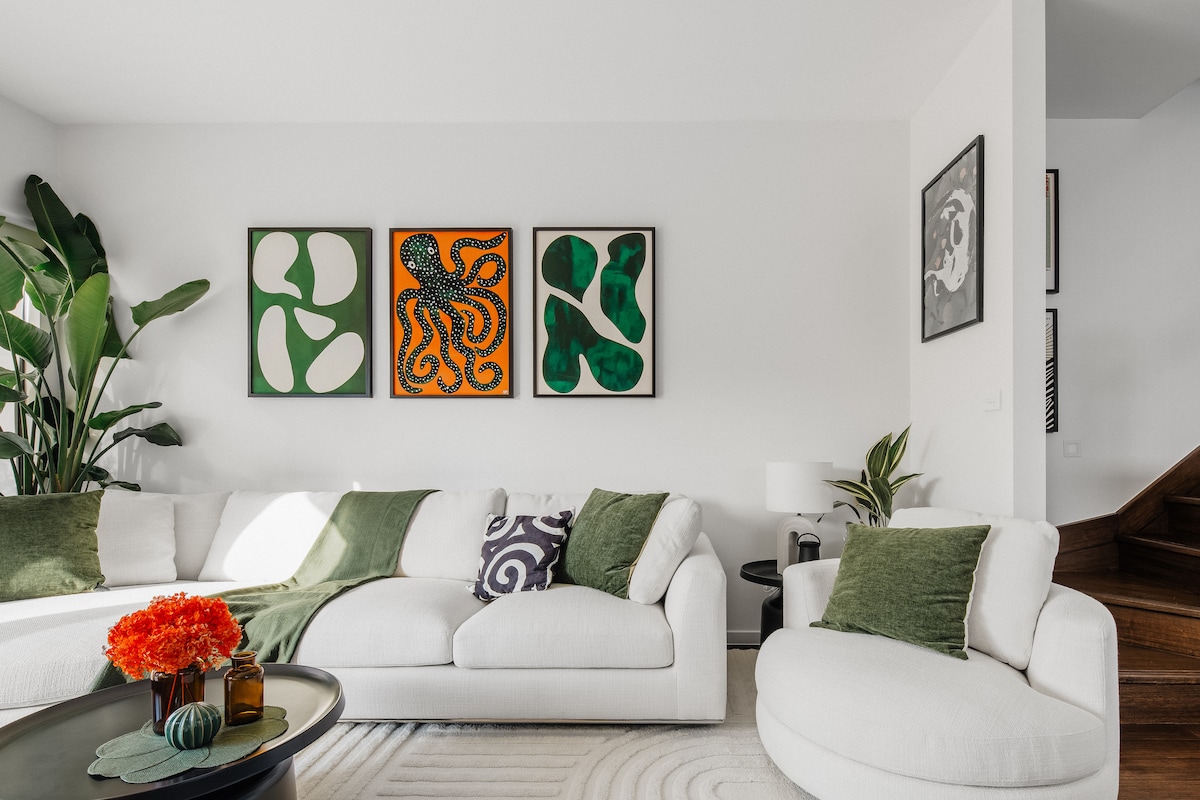 An open living area features a large white sectional sofa adorned with green throw blankets. Abstract art pieces decorate the walls, complementing a potted plant nearby. A round coffee table is arranged with orange flowers and decorative items, creating a balanced and inviting space.