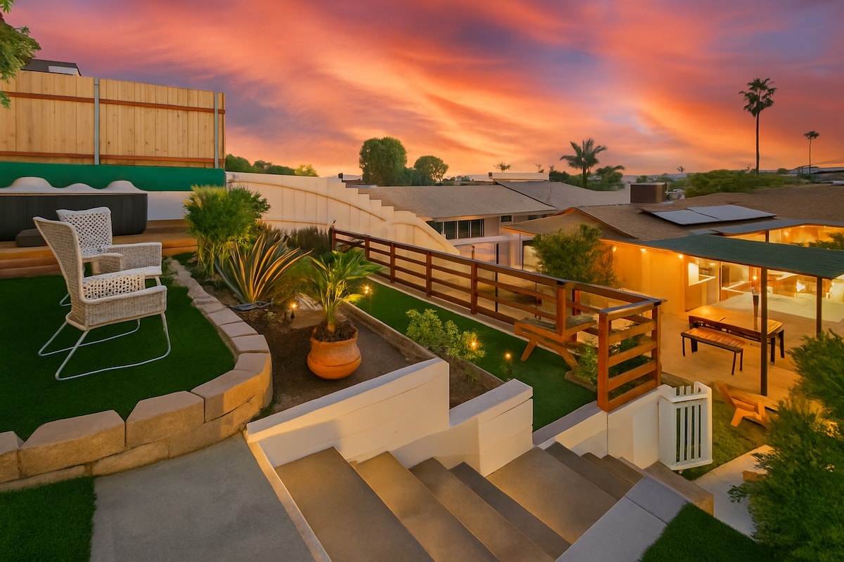 A backyard space is featured, showcasing tiered landscaping with planted greenery and a vibrant sunset sky. Cozy seating is positioned on a patio, and lighting highlights the steps leading down. The area includes a grilling space and a relaxing hot tub in the background.