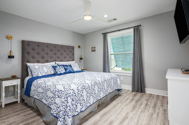 - Пространа брачна спална соба со туфна табла и мека крајбрежна палета на бои
- Сино-бела постелнина нагласена со перница за фрлање на плажа и ѕидни светилки
- Погледот кон прозорецот, вентилаторот на таванот и монтираниот телевизор создаваат простор за опуштање

