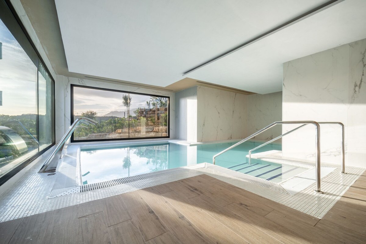 An indoor swimming pool is featured, surrounded by large windows that provide ample natural light and views of the surrounding landscape. The pool is framed by white tiles and has a stainless steel railing leading into the water, enhancing accessibility.