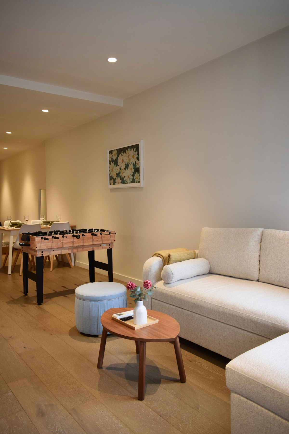 A spacious living area is furnished with a light-colored sofa and a round wooden coffee table. A small side table with flowers adds a touch of decoration. A foosball table is positioned nearby, while a dining setup with a long table is visible in the background.
