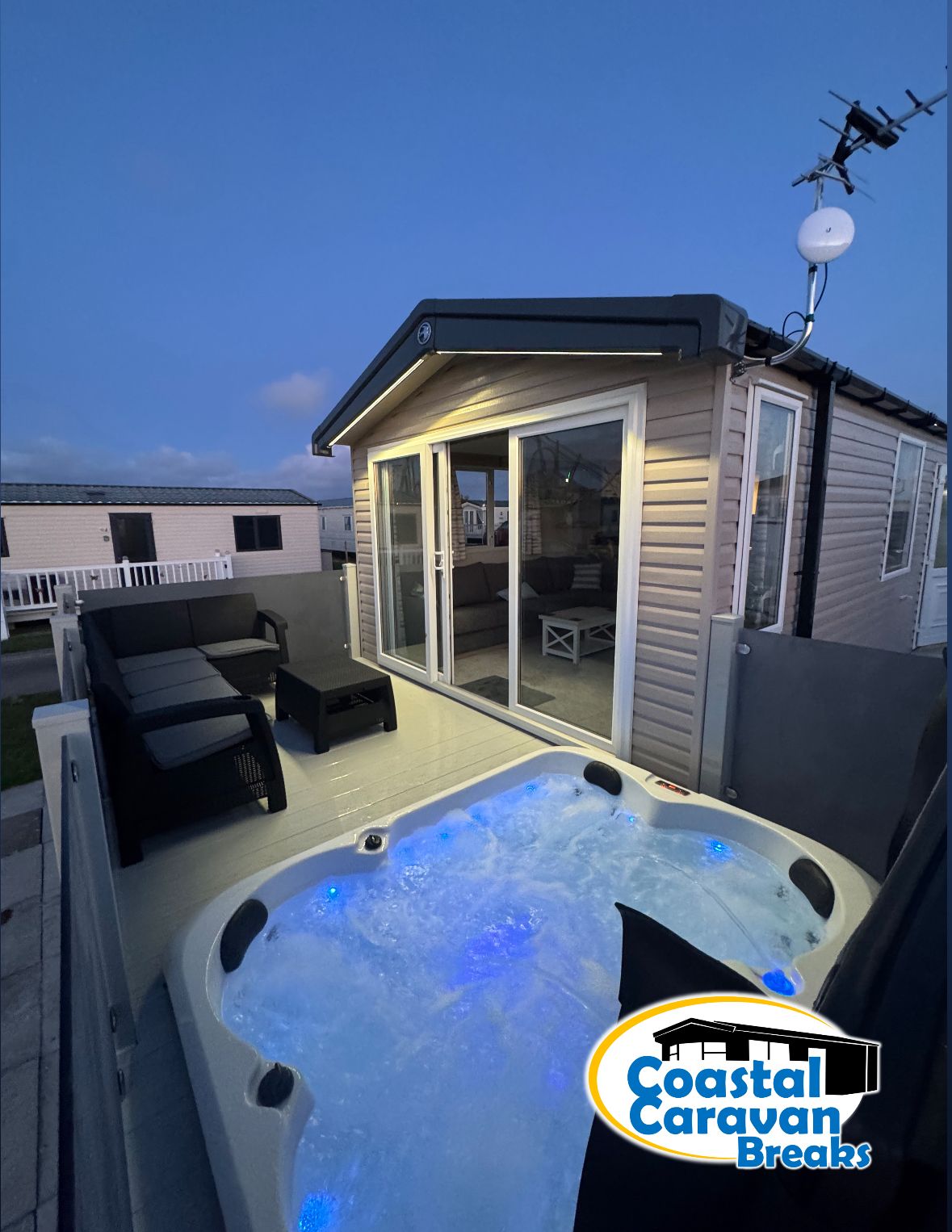 A private outdoor deck is featured, showcasing a hot tub with soothing blue water and LED lighting. Nearby, a modern cabin is visible, characterized by large glass doors that open to a cozy seating area. The scene is set against a twilight sky.