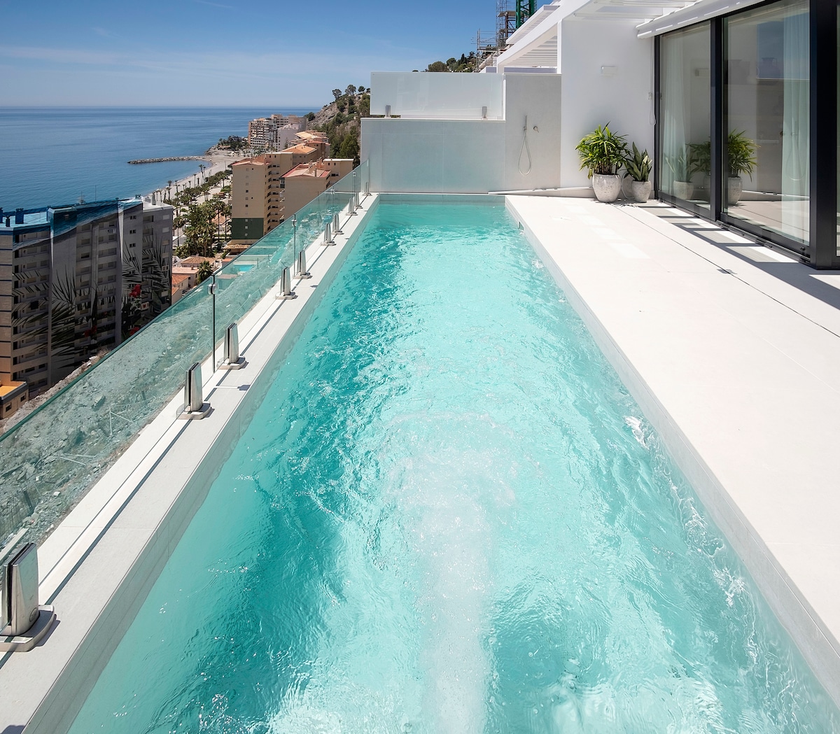 An expansive infinity pool is positioned on a terrace, offering uninterrupted views of the Mediterranean Sea. The pool's clear turquoise water reflects sunlight, creating a serene atmosphere. Glass railings surround the area, enhancing the openness and connection to the surrounding landscape.