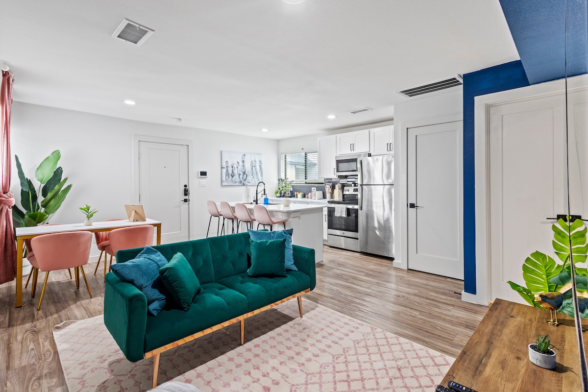 A modern living space features a green sofa placed on a textured rug, complemented by a wooden coffee table. A dining area with pink chairs is visible, alongside a sleek kitchen equipped with stainless steel appliances. Potted plants add a touch of greenery.