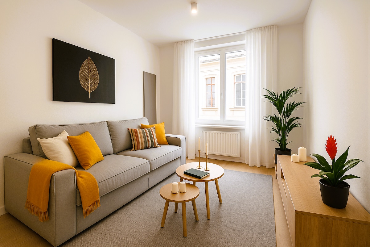 A bright living space is highlighted by a comfortable grey sofa adorned with an orange throw and striped cushions. Minimalist coffee tables are arranged in front, complemented by decorative candles. A plant sits beside the sofa, while soft natural light filters through the large window.