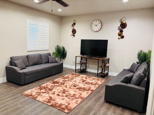 A spacious living area features two gray sofas facing a large television mounted on a stand. A round clock adorns the wall, while decorative wall hangings add a touch of color. A soft, textured area rug in warm tones sits centered on the dark wood floor.
