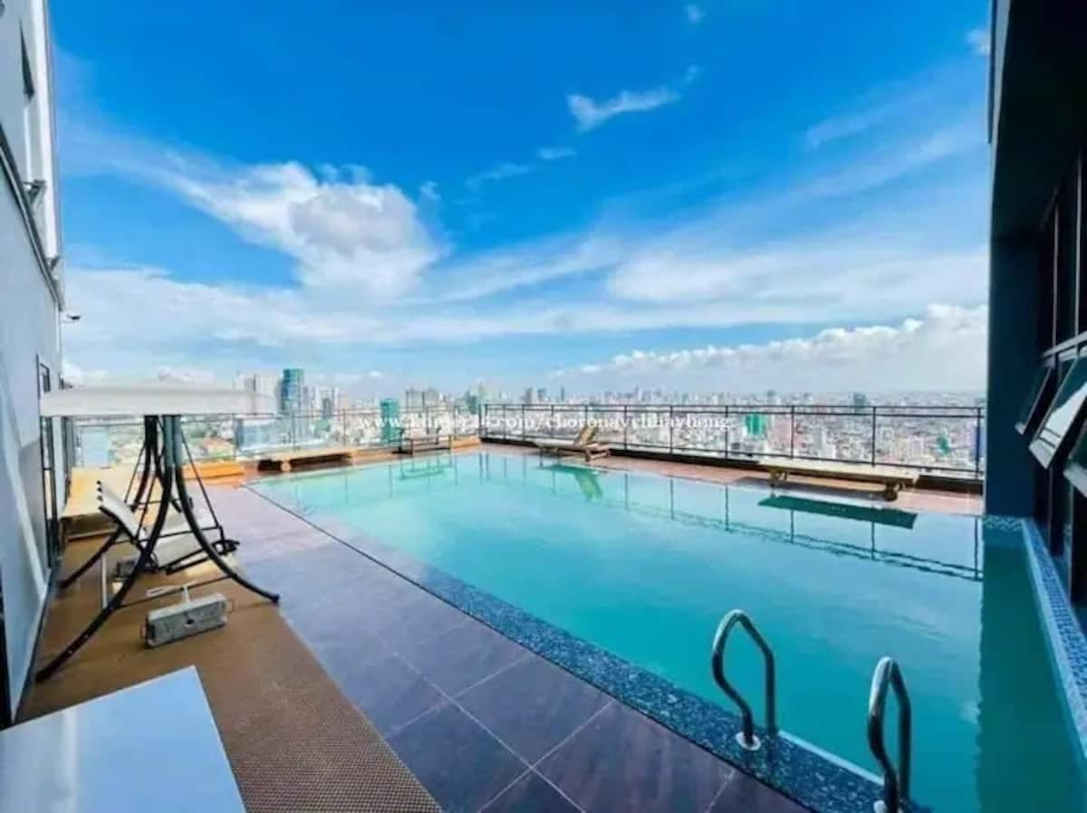 A rooftop pool offers expansive views of the city skyline under a clear blue sky. Lounge chairs are positioned along the poolside, and light reflects off the water, creating a serene atmosphere. The area appears well-maintained and inviting.