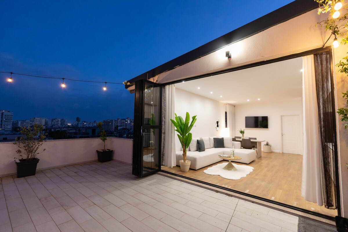 An open living area extends to a spacious outdoor terrace, featuring a light-colored sectional sofa paired with a coffee table. Soft lighting illuminates the space, and potted plants add touches of greenery. The room is framed by large glass doors that create a seamless indoor-outdoor connection.