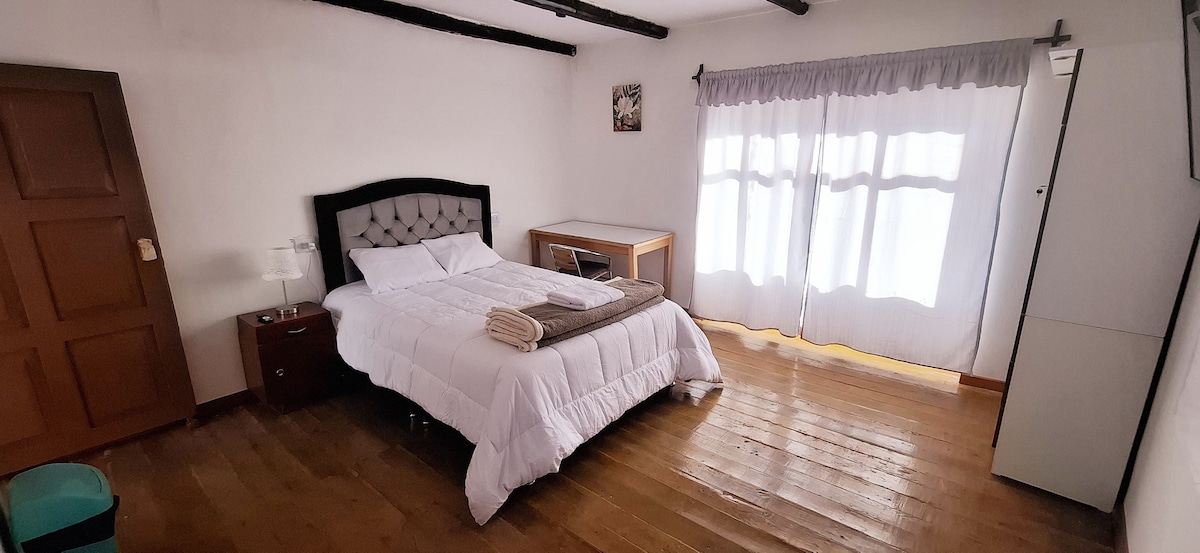 A well-lit bedroom features a spacious bed with crisp white bedding. Natural light filters through sheer curtains, illuminating warm wooden flooring. A bedside table with a lamp and a wooden storage unit are positioned beside the bed, creating an organized space.