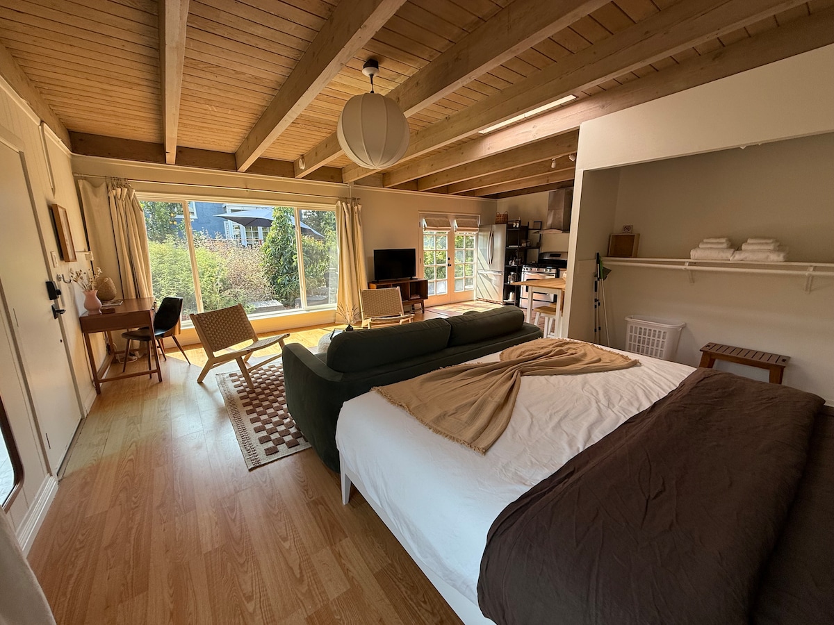 A spacious studio space is showcased, featuring a king-size bed with a soft throw. A large window offers views of the surrounding garden. A desk and chair are positioned near the window, while a cozy couch adds seating. Warm wood accents enhance the inviting atmosphere.