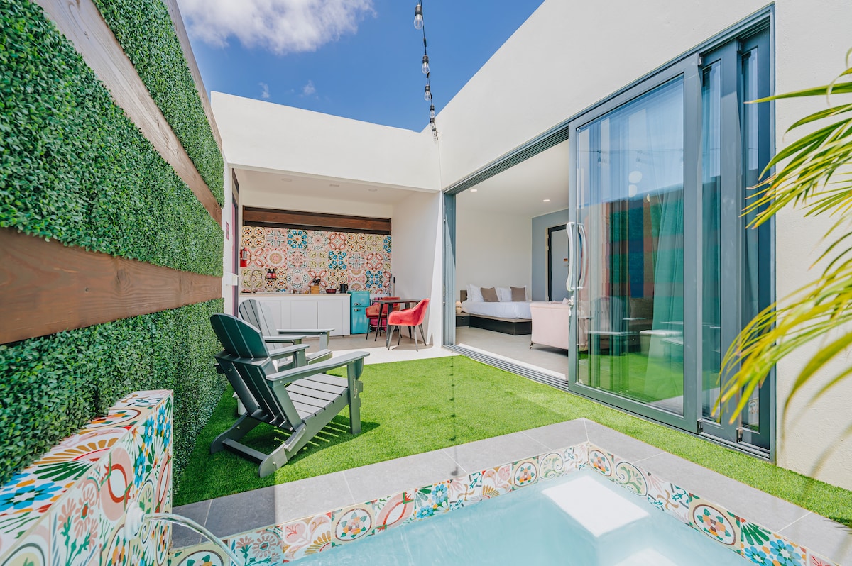 A private garden space is presented, featuring a small plunge pool surrounded by decorative tiles. Green grass covers the ground, complemented by a cozy seating area with two chairs. Large glass sliding doors provide access to a spacious indoor living area, illuminated by natural light.