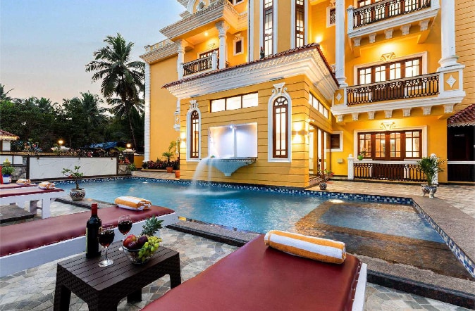 An inviting outdoor area features a private pool with a gentle waterfall, surrounded by sun loungers. Fruit and beverages are displayed on a table nearby. The elegant villa structure rises in the background, adorned with multiple windows and balconies, reflecting golden hues from the setting sun.