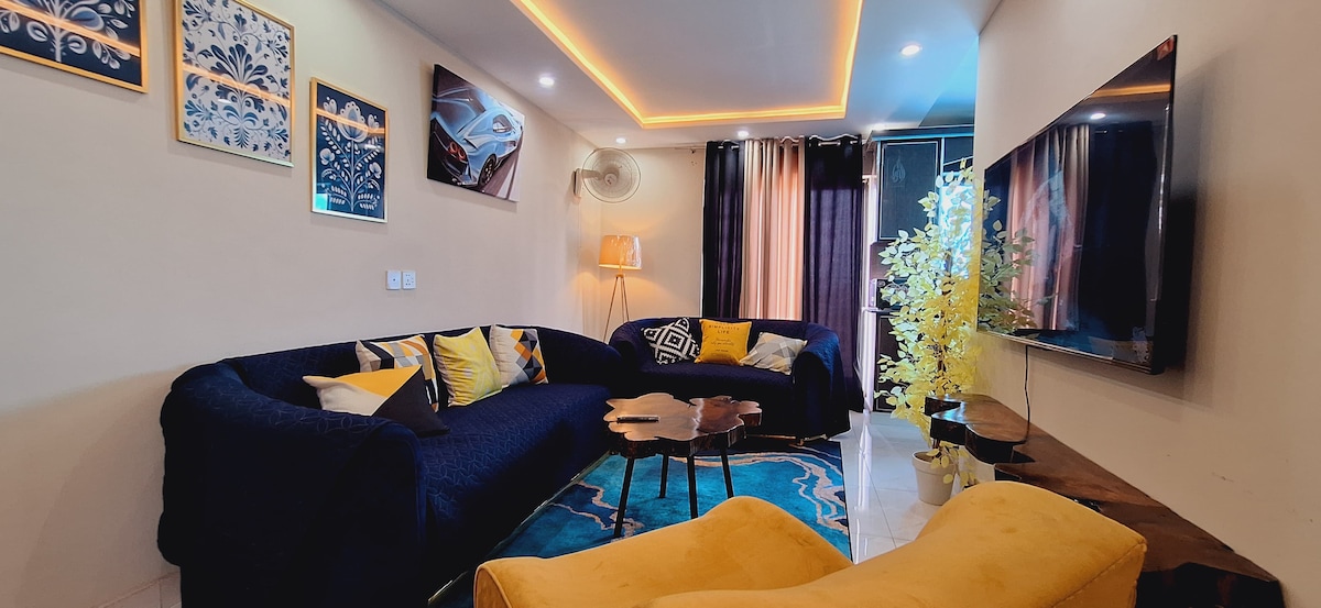 A cozy living space features a dark blue sectional couch adorned with vibrant throw pillows. A small wooden table is positioned in front, and a bright yellow chair adds a pop of color. Ambient lighting and a mounted television enhance the comfortable atmosphere.