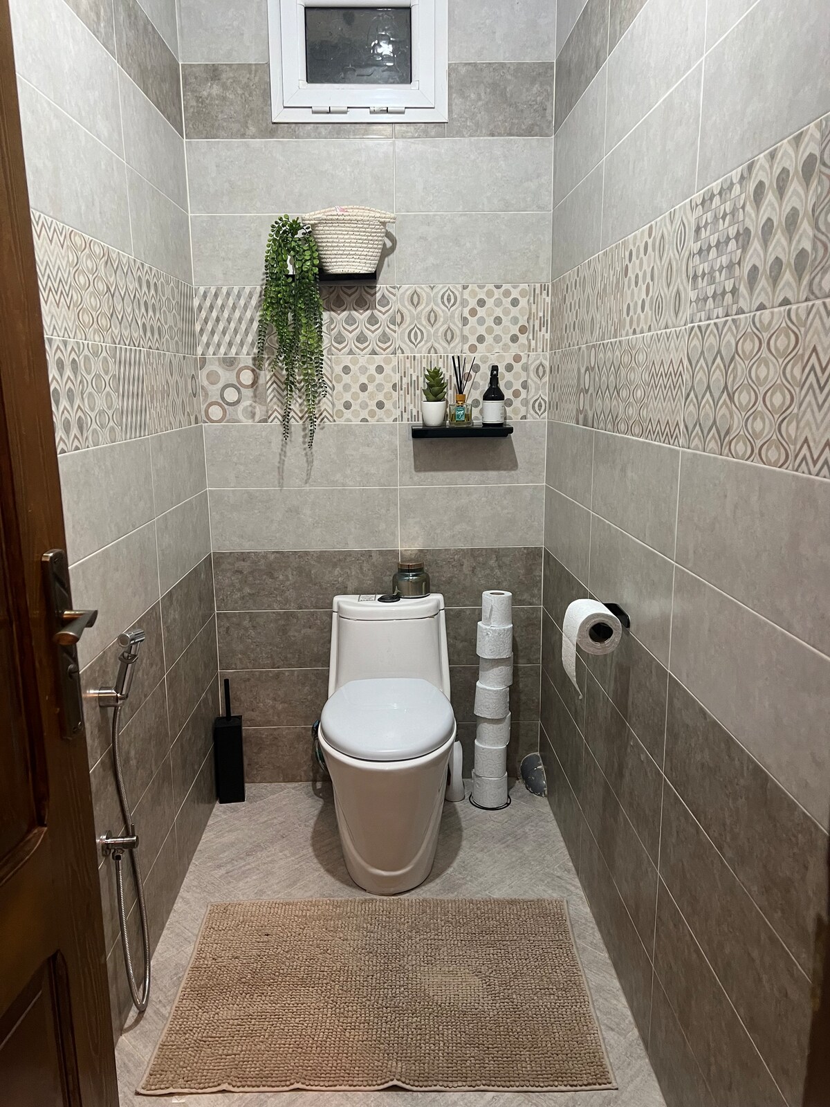 A compact bathroom is presented, featuring neutral tiled walls and a modern toilet. A small shelf holds decorative items and a plant, while essential toiletries are neatly arranged. A bathmat lies on the floor, and a window allows natural light to enter the space.