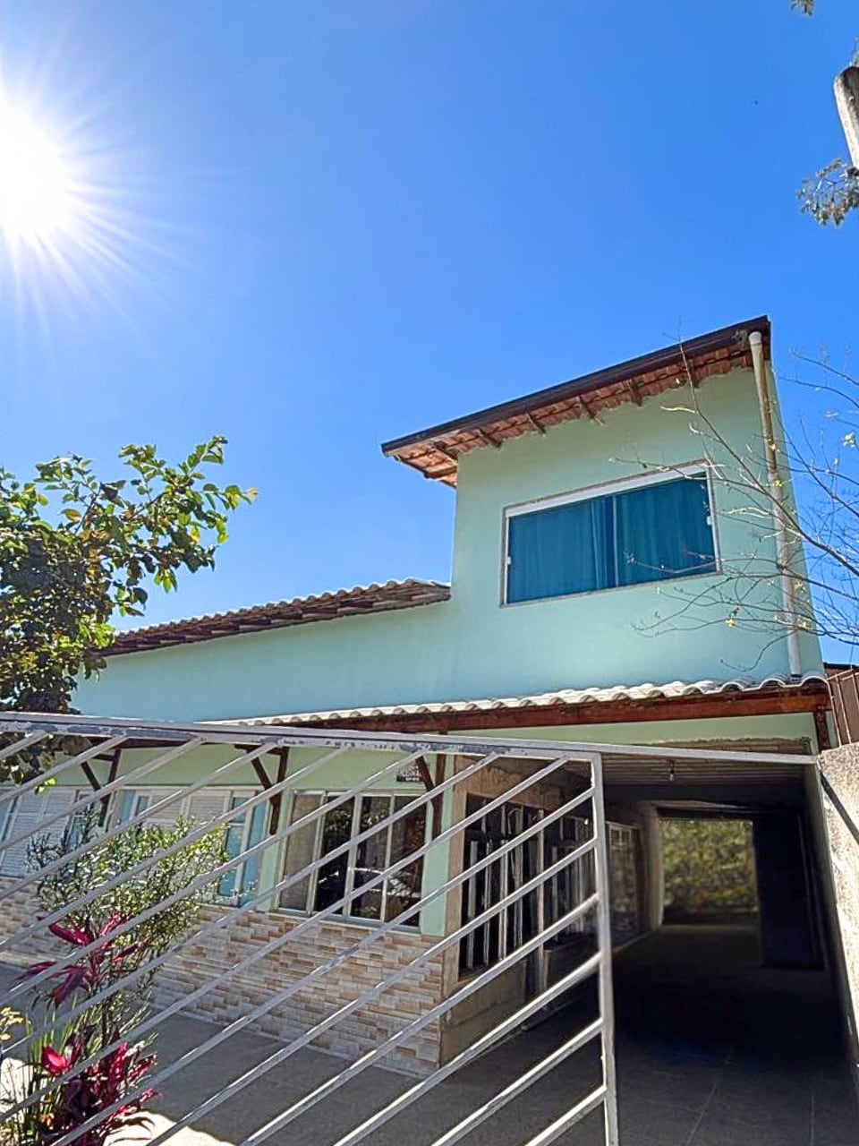 A two-story house is showcased under a clear blue sky, with sunlight reflecting off the surface. A covered parking area is visible, with a gated entrance leading to a spacious patio adorned with greenery.