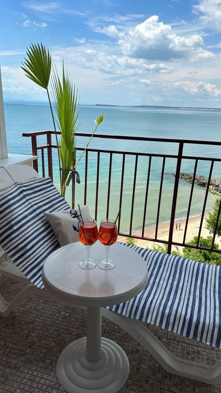 A sunlit terrace offers a relaxing space with a striped lounge chair and a small white table. Two glasses of sparkling drink sit on the table, with a backdrop of the sea and distant sandy beach visible in the horizon.