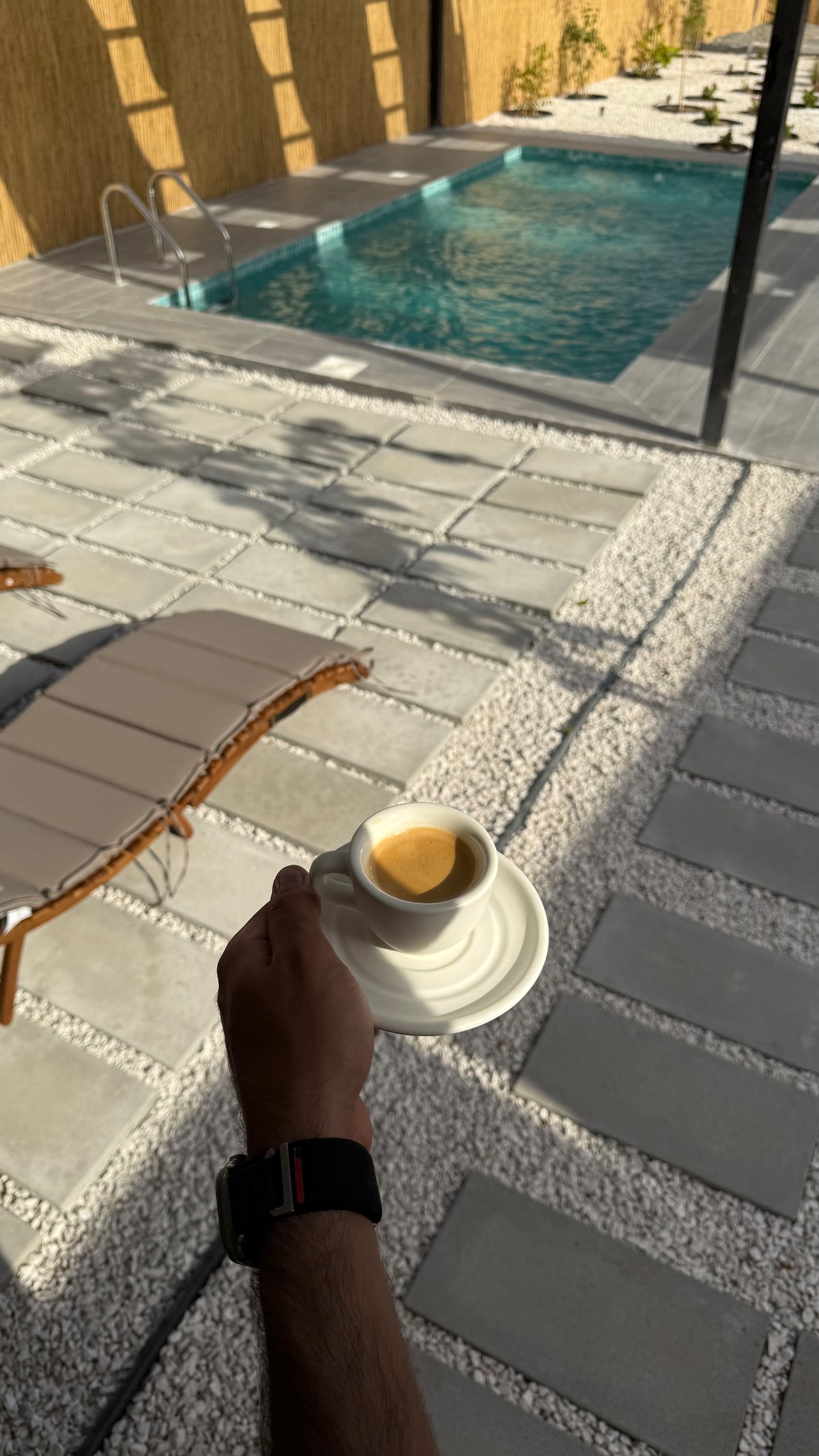 A hand holds a small cup of coffee, accompanied by a white saucer. In the background, a swimming pool is visible, surrounded by a light-colored stone patio and neatly arranged small plants. A sun lounger rests nearby, complementing the relaxing atmosphere.