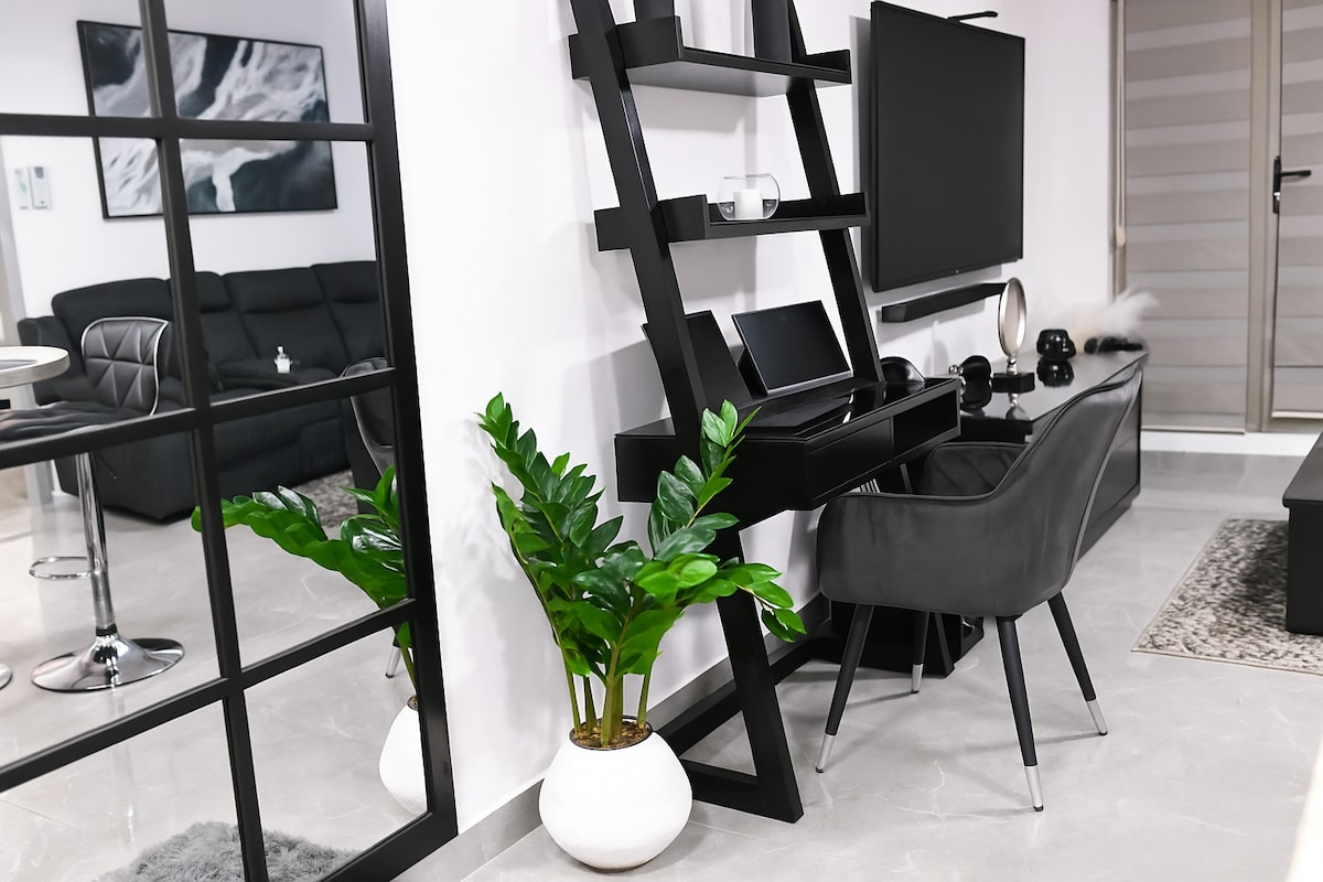 A modern living space features a sleek black bookshelf against a white wall, complemented by a stylish chair with tapered legs. A flat-screen TV is mounted nearby, while a small plant in a white pot adds a touch of greenery to the ambiance.