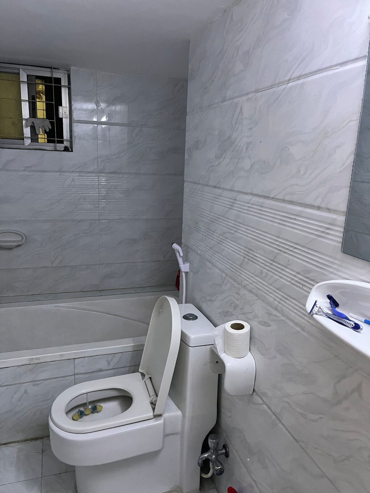A clean, modern bathroom features light-colored tiled walls and a spacious bathtub. A white toilet is positioned adjacent to a small washbasin that holds toiletries. Natural light enters through a window, enhancing the room's serene atmosphere.