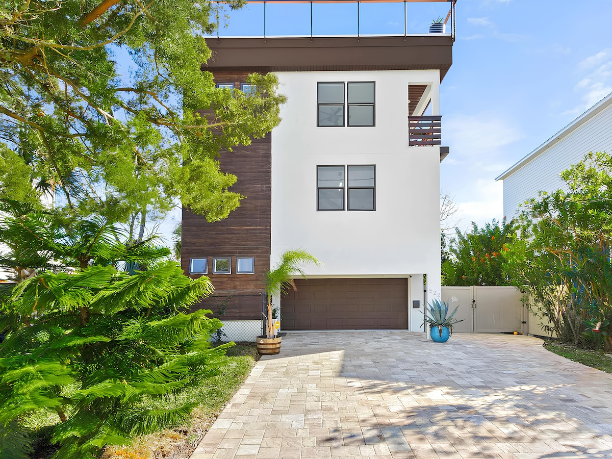 The exterior of a three-story home is displayed, featuring a modern design with a clean white facade and natural wood accents. A spacious driveway is visible, surrounded by lush greenery, and large windows allow for ample natural light. A balcony extends from the upper floor.