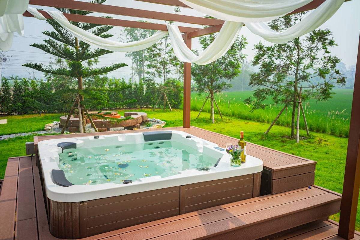 An outdoor jacuzzi designed for four individuals is nestled beneath a shaded pergola. Surrounding greenery and decorative landscaping create a serene environment. A small table with refreshments sits nearby, enhancing the inviting outdoor space.