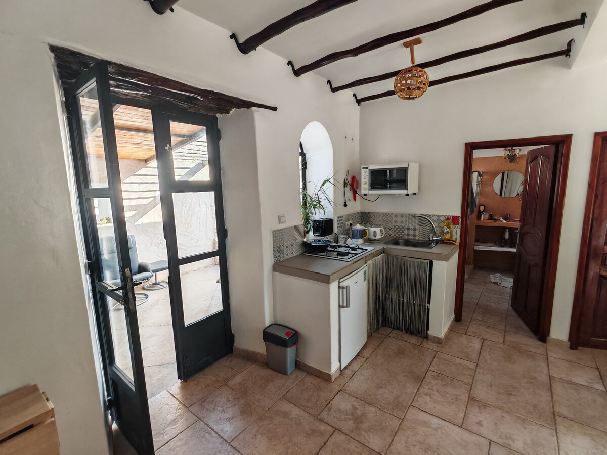 A functional kitchen area is visible, featuring wood beam ceilings and tiled flooring. Appliances include a microwave and stovetop, with a small table set against the wall. Natural light enters through a glass door that leads to an outdoor space.